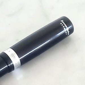 K&ouml;nnte beinhalten: Nahaufnahme eines schwarzen Stifts mit gl&auml;nzendem Finish. Der Stift hat ein silbernes Band mit dem Aufdruck "GERMANY". Auf der Kappe des Stifts stehen die W&ouml;rter "Kaweco" und "GERMANY" in Wei&szlig;.
