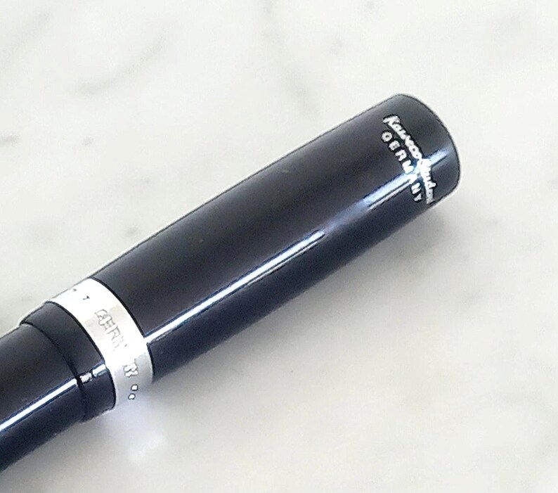 K&ouml;nnte beinhalten: Nahaufnahme eines schwarzen Stifts mit gl&auml;nzendem Finish. Der Stift hat ein silbernes Band mit dem Aufdruck "GERMANY". Auf der Kappe des Stifts stehen die W&ouml;rter "Kaweco" und "GERMANY" in Wei&szlig;.