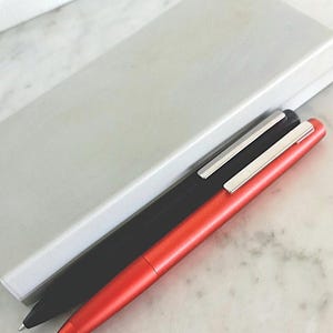 May include: Two sleek pens, one black and one red, with silver accents, rest beside a light gray pen case. The pens have a minimalist design and are set against a marble surface.