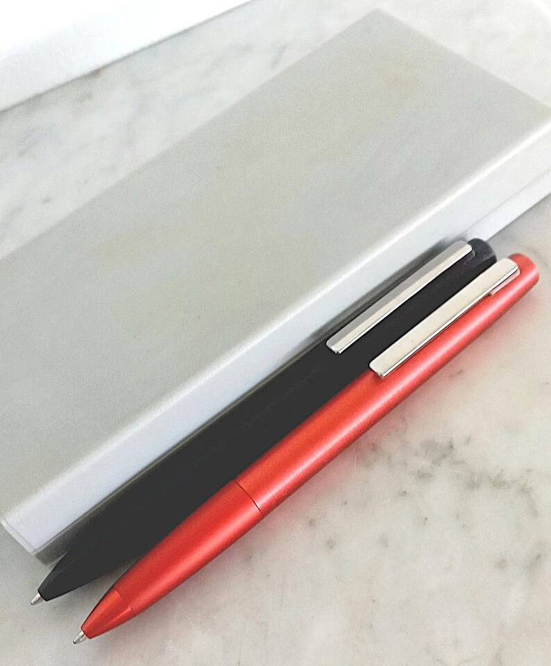 May include: Two sleek pens, one black and one red, with silver accents, rest beside a light gray pen case. The pens have a minimalist design and are set against a marble surface.