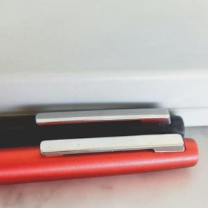 May include: Close-up of two pens, one red and one black, with silver-colored clips. The pens are lying on top of each other, with the red pen in the foreground. The background is a white wall.