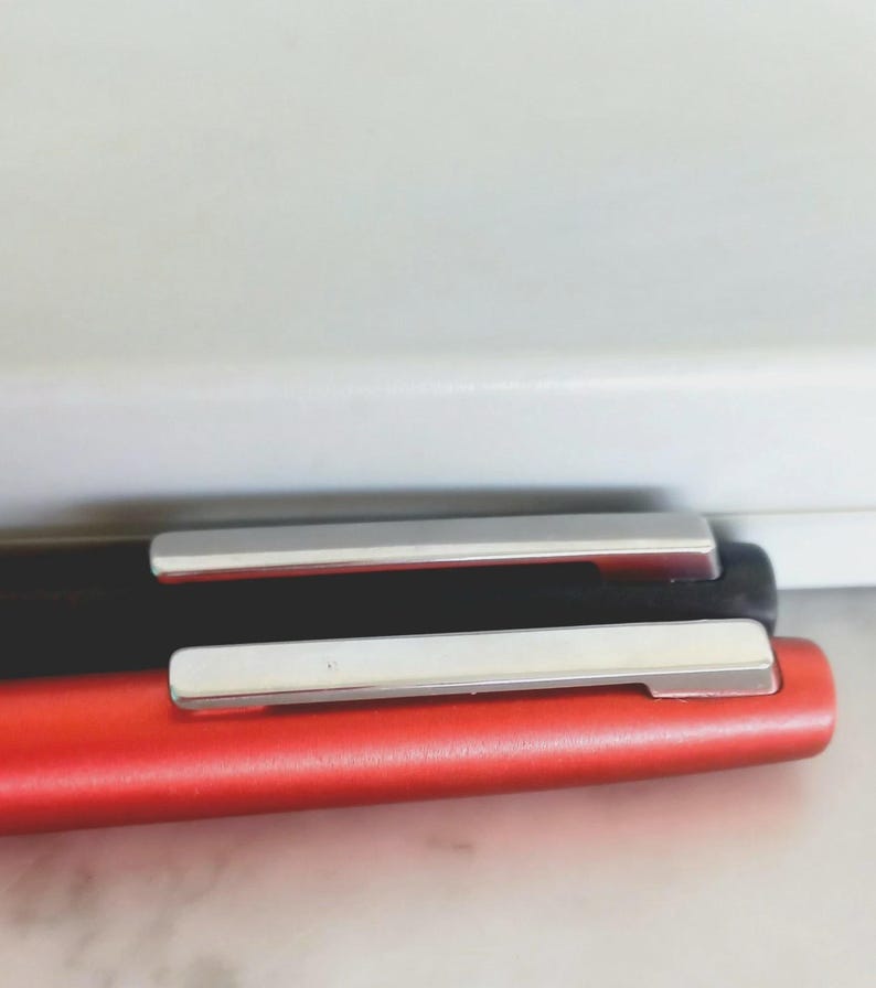 May include: Close-up of two pens, one red and one black, with silver-colored clips. The pens are lying on top of each other, with the red pen in the foreground. The background is a white wall.