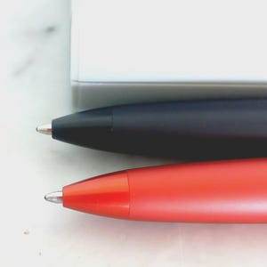 May include: Close-up of two pens. One pen is black, and the other is red. Both pens have a silver tip and a sleek, modern design. The pens are positioned horizontally against a white background.