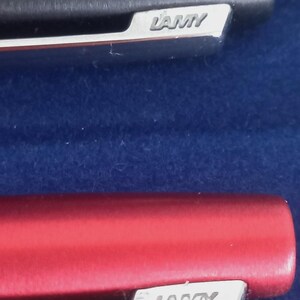 May include: Close-up of two Lamy pens. One pen is black, the other is red. Both pens have a silver clip with the Lamy logo. The pens are set against a dark blue background.