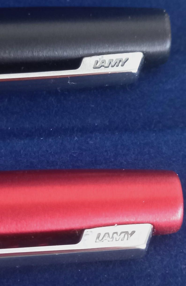 May include: Close-up of two Lamy pens. One pen is black, the other is red. Both pens have a silver clip with the Lamy logo. The pens are set against a dark blue background.