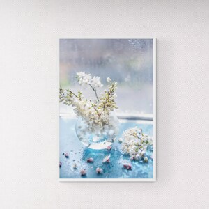 Puede incluir: Un jarrón de vidrio lleno de flores blancas se encuentra sobre una superficie azul con gotas de agua. El jarrón es redondo y transparente, y las flores son delicadas y suaves. El fondo es una imagen borrosa de una ventana con gotas de lluvia.