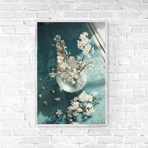 Puede incluir: Un jarrón de vidrio lleno de flores de cerezo blancas se encuentra sobre una superficie azul turquesa. El jarrón es redondo y transparente, y las flores son delicadas y fragantes.