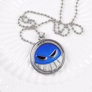 Może przedstawiać: Srebrny naszyjnik z okrągłą zawieszką przedstawiającą niebieską twarz z czarnymi oczami i białym uśmiechem. Zawieszka jest na srebrnym łańcuszku kulkowym, prezentowana na białym talerzu.