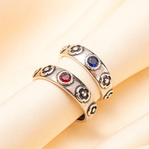 Può includere: Due anelli d'argento con motivi floreali e pietre preziose rosse e blu. Gli anelli sono su uno sfondo di tessuto color crema.