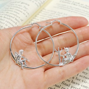 May include: Silver hoop earrings with a cherub and a devil charm. The earrings are shown on a hand against a book background.