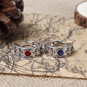 Può includere: Due anelli d'argento con disegni decorativi. Un anello ha una pietra preziosa rossa, l'altro ha una pietra preziosa blu.