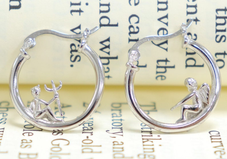 May include: A pair of silver hoop earrings with a small angel and a small devil charm hanging from each hoop.