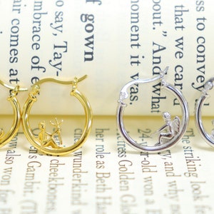 May include: Two pairs of hoop earrings with a small, detailed design inside the hoop. One pair is gold with a fairy and a deer, the other is silver with a fairy and a trident.