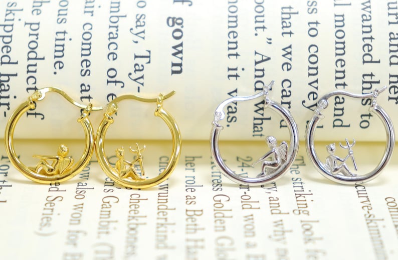 May include: Two pairs of hoop earrings with a small, detailed design inside the hoop. One pair is gold with a fairy and a deer, the other is silver with a fairy and a trident.