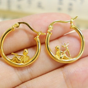 May include: Gold hoop earrings with a small angel figure design. The angel is sitting with wings outstretched and holding a musical instrument.