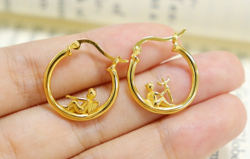 May include: Gold hoop earrings with a small angel figure design. The angel is sitting with wings outstretched and holding a musical instrument.