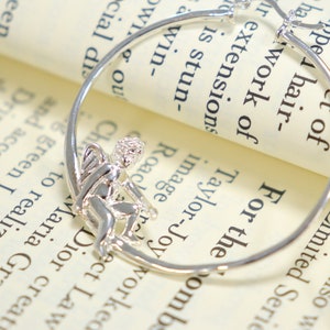 May include: Silver hoop earrings with a small angel charm hanging from one hoop. The earrings are on a white surface with text.