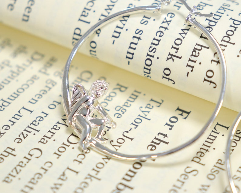 May include: Silver hoop earrings with a small angel charm hanging from one hoop. The earrings are on a white surface with text.