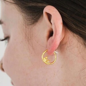 May include: Gold hoop earrings with a small figure of a person sitting inside the hoop.