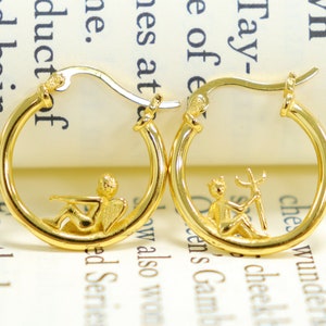 May include: A pair of gold hoop earrings with a small angel and a small figure holding a trident hanging from each hoop.