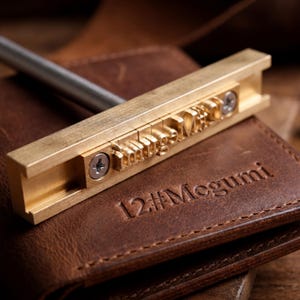 May include: A brass branding iron with the text "handmade" is positioned over a brown leather wallet. The wallet has the name "Megumi" and the number "12" embossed on it. The branding iron has a silver handle.
