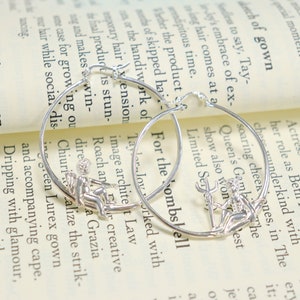 May include: A pair of silver hoop earrings with a small, detailed charm of a woman holding a trident. The earrings are on a white background with text.