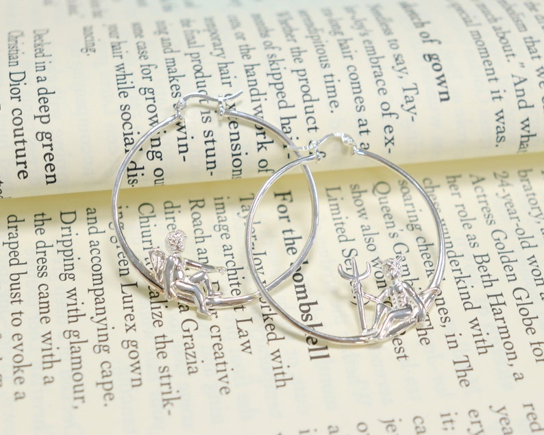 May include: A pair of silver hoop earrings with a small, detailed charm of a woman holding a trident. The earrings are on a white background with text.