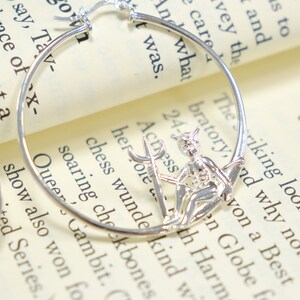 May include: Silver hoop earrings with a small silver charm of a winged figure holding a trident. The charm is attached to the hoop earring.