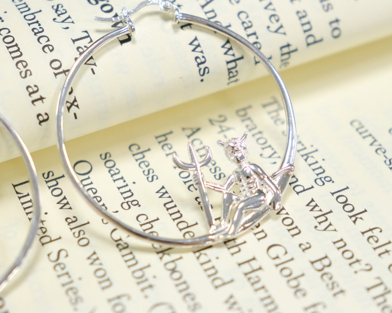 May include: Silver hoop earrings with a small silver charm of a winged figure holding a trident. The charm is attached to the hoop earring.