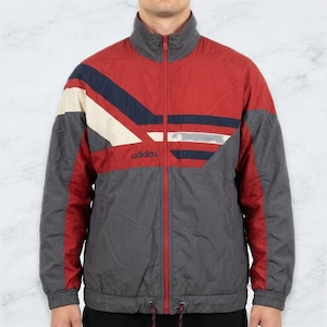 May include: A red, dark gray, and cream-colored Adidas track jacket featuring a high collar, a full-length front zipper, and long sleeves. The jacket displays a geometric color-blocked pattern across the chest and shoulders with the Adidas logo.