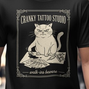 May include: Black t-shirt featuring a grumpy cat illustration with the text "Cranky Tattoo Studio - walk-ins beware." The cat is depicted with a stern expression, surrounded by tattoo-related items.