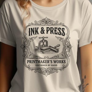 May include: Cream-colored t-shirt featuring a black and white graphic design. The design includes the text "INK & PRESS" above an illustration of a printing press and the text "PRINTMAKER'S WORKS" below. The shirt is a classic crew neck.