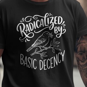 以下が含まれることがあります： 「Radicalized by Basic Decency」の白い文字がプリントされた黒いTシャツ。テキストの下には、枝にとまっている鳥の精巧なイラストが描かれています。Tシャツは柔らかい素材で作られています。