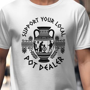 May include: A white short-sleeved t-shirt featuring a black graphic of an ancient Greek-style amphora vase. The text "SUPPORT YOUR LOCAL POT DEALER" is printed in a circular pattern around the central illustration of a potter at a wheel.