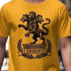 以下が含まれることがあります： マスタードイエローの半袖Tシャツ。台座の上に立つ3つの頭を持つ犬ケルベロスの黒いグラフィックプリントが施されています。デザインには月桂樹の冠の縁取りと、生き物の下のバナーの中央にCERBERUSというテキストが含まれています。