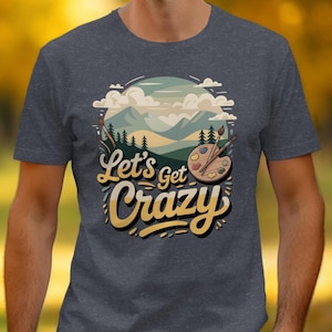 Puede incluir: Camiseta gris oscuro con un gráfico colorido. El diseño presenta un paisaje montañoso, una paleta de pintura y el texto "Let's get Crazy" en una fuente lúdica. La camiseta es de manga corta y tiene cuello redondo.