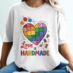 May include: A white t-shirt featuring a colourful patchwork heart graphic made of various fabric patterns. Surrounding the heart are illustrations of yarn balls, sewing spools, and needles. The text reads Love IS HANDMADE in colourful letters.