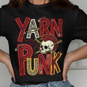 May include: Black t-shirt with the words "YARN PUNK" in red, white, and yellow, featuring a skull with a mohawk and knitting needles. The design incorporates yarn and safety pins.