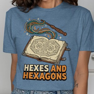 May include: Blue t-shirt with a graphic of an open book, a wand, and the text "HEXES AND HEXAGONS". The book has detailed illustrations, and the wand emits colorful strands. The design includes stars and crescent moons.