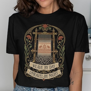 May include: A black short-sleeve t-shirt featuring a graphic of a wooden loom with a woven tapestry depicting two birds and a tree. The design includes floral accents and a banner with the text WEAVE BY DAY UNRAVEL BY NIGHT in gold.