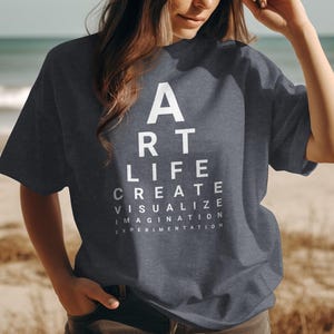 May include: Dark gray t-shirt with white text arranged like an eye chart: ART, LIFE, CREATE, VISUALIZE, IMAGINATION, EXPERIMENTATION. The shirt is made of a soft material and is suitable for casual wear. The background is a beach scene.