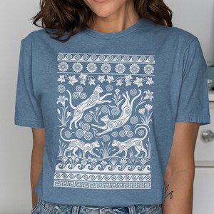 Könnte beinhalten: Ein kurzärmeliges T-Shirt in Heideblau mit einem weißen Grafikdruck von vier Katzen, umgeben von Blumenmotiven, Spiralmustern und geometrischen Bordüren. Das Design enthält stilisierte Blätter, Blüten und wellenförmige Linien in einem Muster.