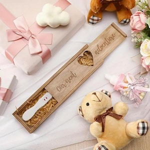 Op de afbeelding: Een houten doos met de inscriptie "Our next adventure", met een positieve zwangerschapstest. De doos is open en onthult de test op stro. De afbeelding bevat een teddybeer, cadeaus en een babyfles.