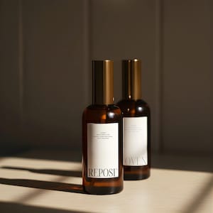 May include: Two amber glass spray bottles with gold-colored caps and white labels. The front bottle has the word "REPOSE" printed on it. The other bottle has the word "LOVEN" printed on it. The bottles are on a light-colored surface.