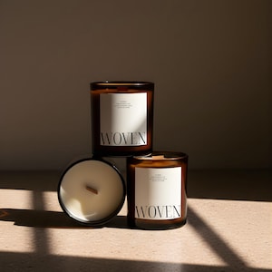 May include: Three brown glass candles with white labels that read "WOVEN". The candles have a white wax and a wooden wick. The candles are arranged in a triangular shape, with one candle on its side.