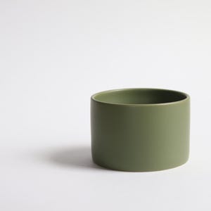 Puede incluir: Un recipiente cilíndrico de cerámica verde oliva. El recipiente tiene un acabado liso y mate y un diseño sencillo y moderno. El recipiente tiene aproximadamente 10 cm de diámetro y 7,6 cm de altura. Está sobre un fondo blanco liso.