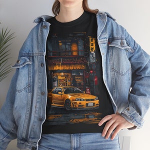 Könnte beinhalten: Ein schwarzes T-Shirt mit einem Aufdruck eines gelben Autos, das vor einem japanischen Geschäft mit dem Text "青春時代" in japanischer Sprache geparkt ist. Das Geschäft ist mit warmen gelben Lichtern beleuchtet und bietet eine Auswahl an Waren zum Verkauf.
