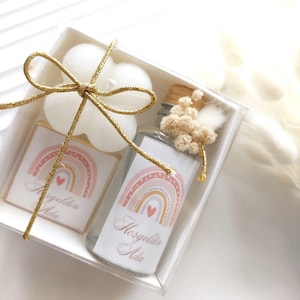 Puede incluir: Una caja de regalo transparente con una cinta dorada atada alrededor. Dentro de la caja hay dos velas blancas, una pequeña botella de líquido transparente con una etiqueta que dice "Bienvenido Ada" y un diseño de arcoíris, y una pequeña tarjeta blanca con el mismo diseño de arcoíris y el texto "Bienvenido Ada".