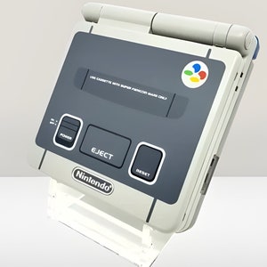 Puede incluir: Una consola de videojuegos portátil blanca y gris con una ranura para cartuchos gris y botones etiquetados como "EJECT", "POWER" y "RESET". La consola tiene un logotipo de Nintendo y el texto "USE CARTRIDGE WITH SUPER Famicom/SUPER NES/FAMILY  COM ONLY?" impreso en ella.