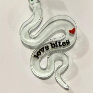 May include: Clear acrylic snake-shaped charm with the words "love bites" in black. The charm has a red heart at the top and a small red heart on the snake's body. The snake has green eyes.